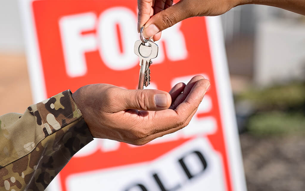 A hand of a military member receiving a key to a home in front of a for sale sign.