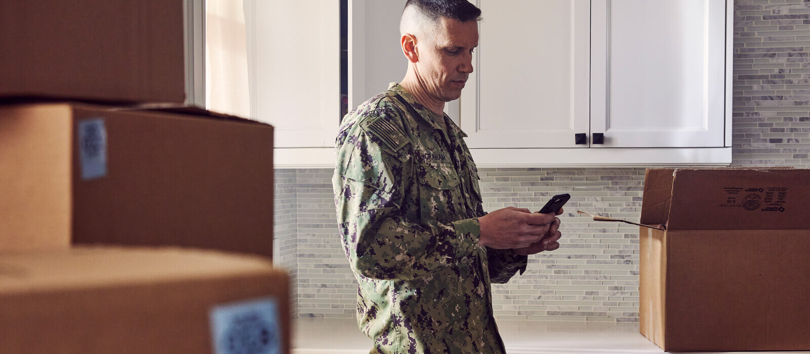 Servicemember texting on a phone surrounded by packing boxes.