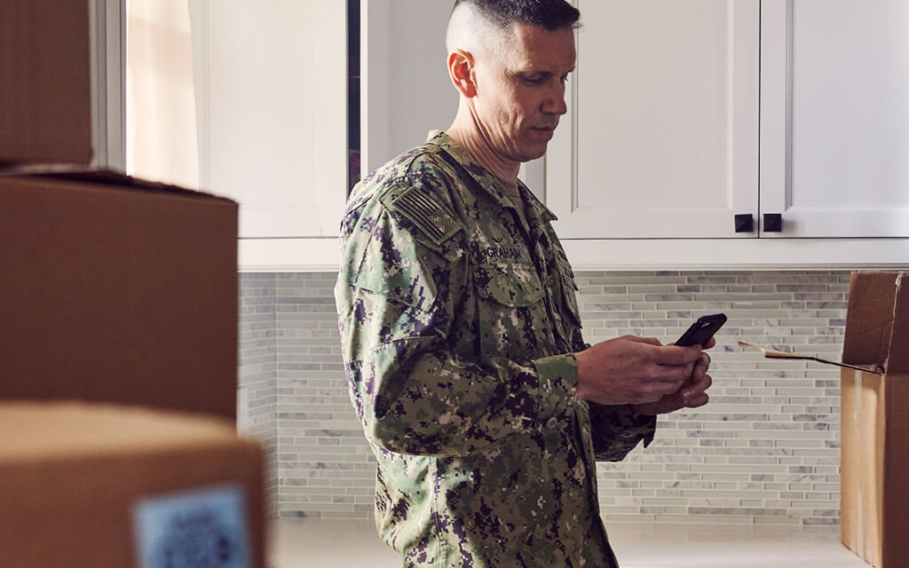 An image of a service member surrounded by moving boxes looking at his home mortgage on his phone.
