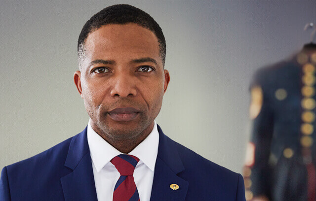 A man in a suit, looking forward intently with a military uniform behind him